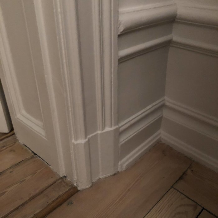 Hörnet av ett rum med en närbild av ett vitt dörrfoder och en matchande fodersockel vid ett trägolv.
