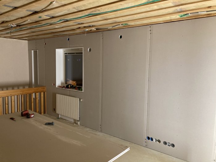 Nyinstallerade gipsskivor i ett vardagsrum under renovering, med synliga ledningar och rörinstallationer.