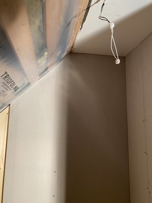 Hörna av ett rum under renovering med gipsskivor monterade på väggarna och hängande elkabel från taket.