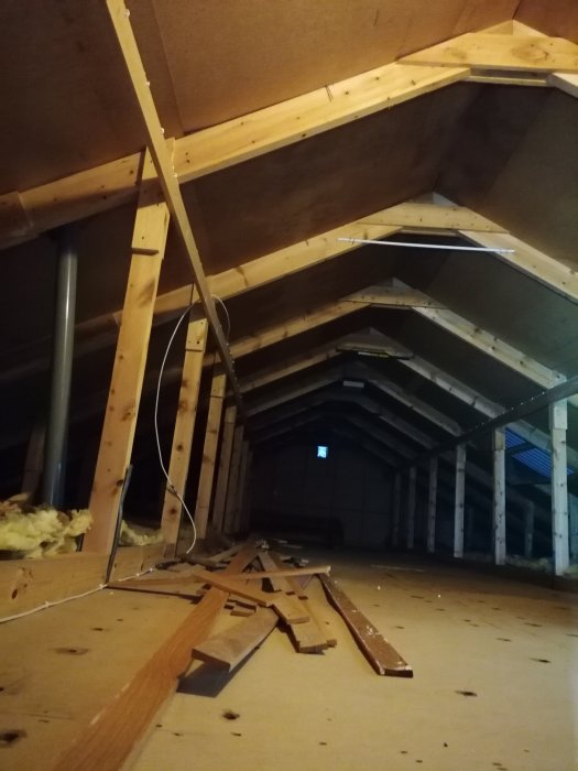 Regelverk och träbjälkar i ett vindutrymme med utspridda brädor och isoleringsmaterial på golvet.