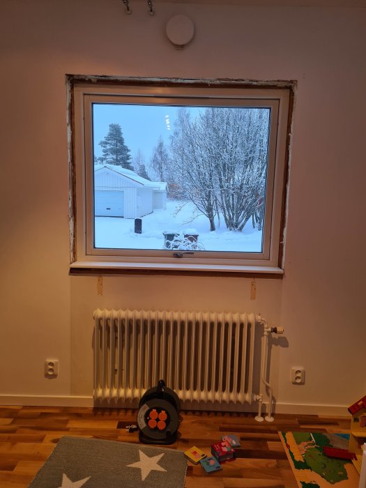 Fönsterutsikt med snö, barnrum redo för renovering med radiator och eluttag, leksaker på golvet.