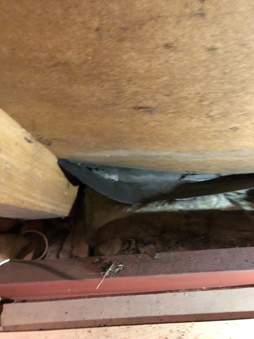 Överblick av en vind med exponerad isolering och delvis släppt vindpapp, med synliga träbjälkar och grus.