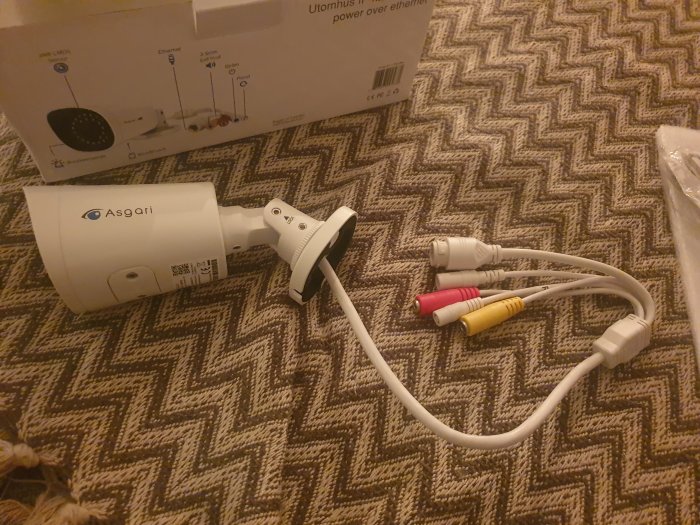 Säkerhetskamera med monteringsarm och utstickande nätkabel samt tre AV-kontakter bredvid dess kartongförpackning.