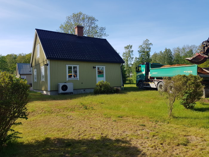 Gult hus med svart tak och avloppsarbete utförs med en grävmaskin och lastbil synlig på gården.