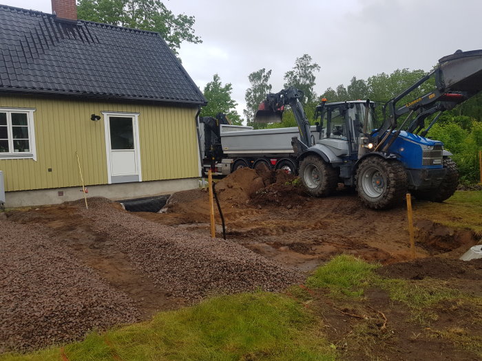 Grävmaskin och lastbil vid markarbete för avlopp utanför ett gult hus med svart tak.