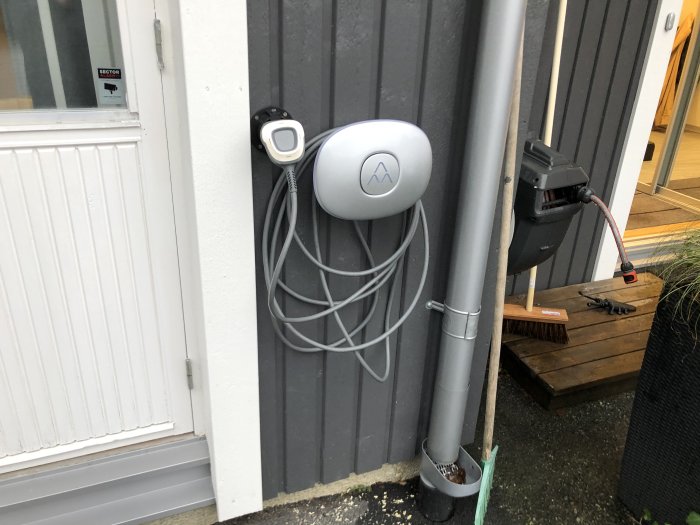 Laddbox för elbil installerad på en byggnads vägg bredvid en hängränna och ett ställ med handverktyg.