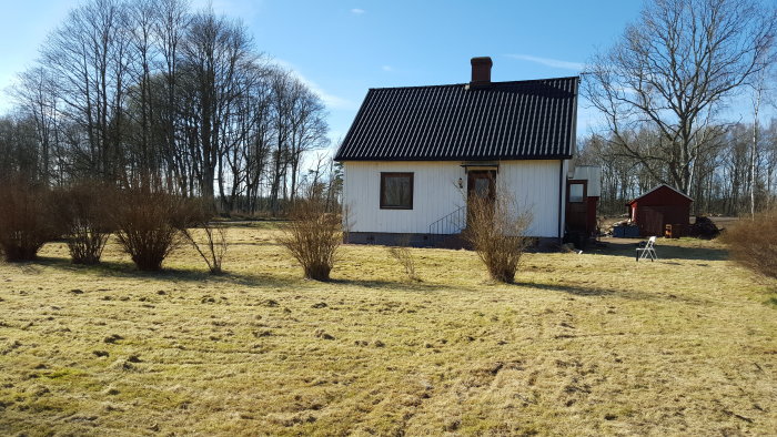 Ett hus med sliten gavel och presenning mot en bakgrund av träd och en gräsmatta med trädgårdsredskap.