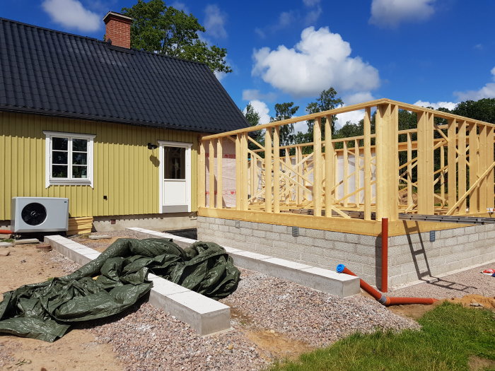 Golvbjälklag och väggstommar under uppbyggnad vid en husutvidgning med presenning på marken.