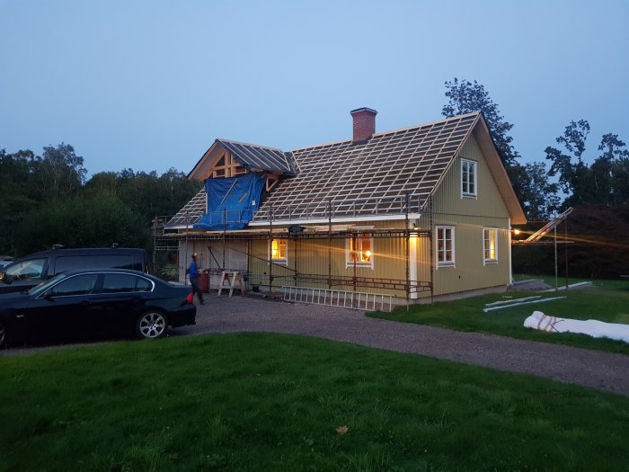 Hus under renovering med delvis lagt tegeltak och blå presenning, i skymningen med tända lampor.