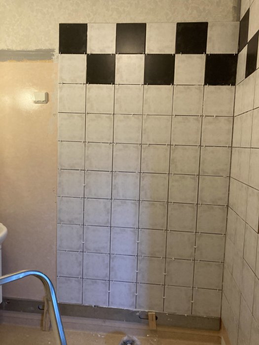 Delvis kaklad vägg med svarta och vita plattor och ett synligt vägguttag, plattorna är ej helt monterade.