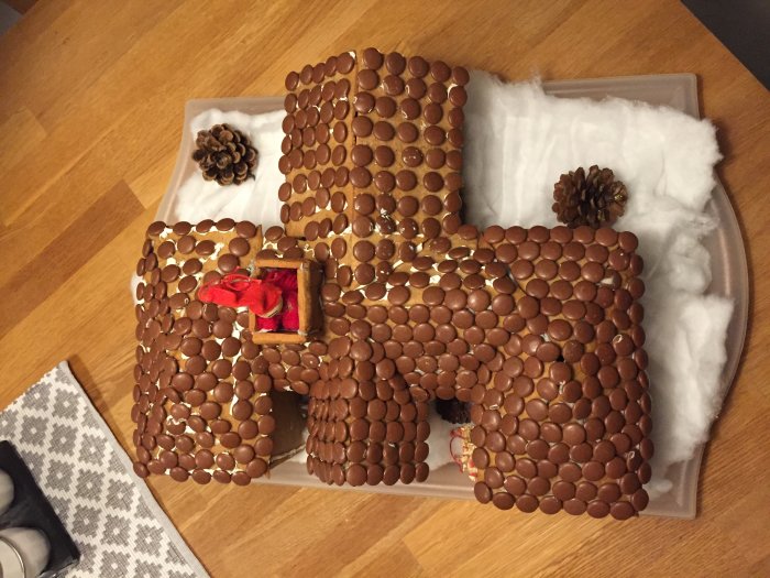 Kreativ miniatyrkopia av ett hus gjord av pepparkakor och chokladpraliner, dekorerad med en småfigur och kottar.