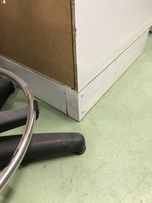 Del av vit skåpstomme med sockel på grönt golv bredvid svart kontorsstolsdel.
