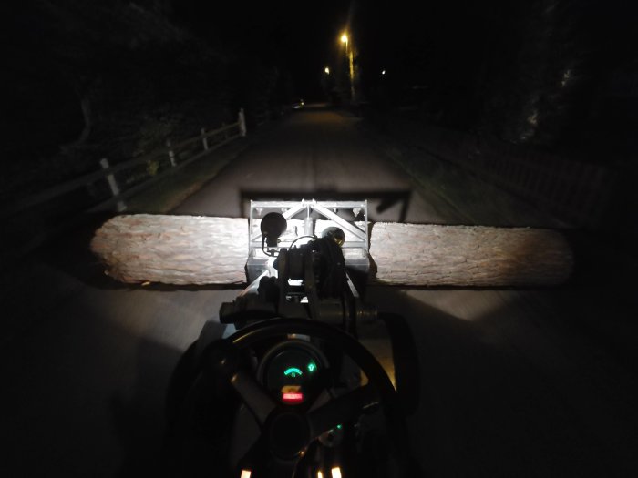 Minilastare nattetid transporterar en stor stock längs en mörk väg, belyst av lastarens strålkastare.