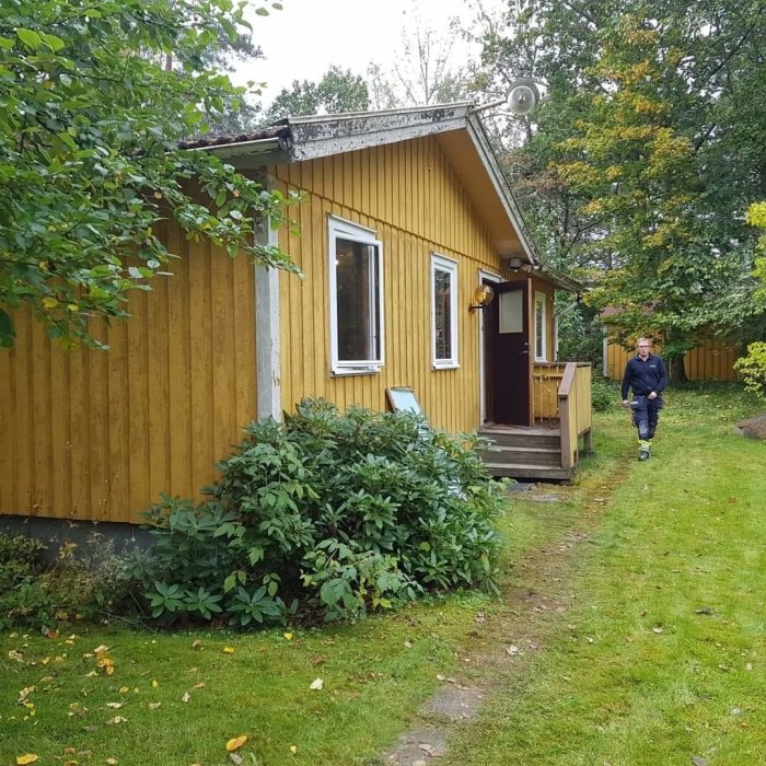 Gul trävilla från 1976 med entrétrappa och grönska, person går i trädgården.