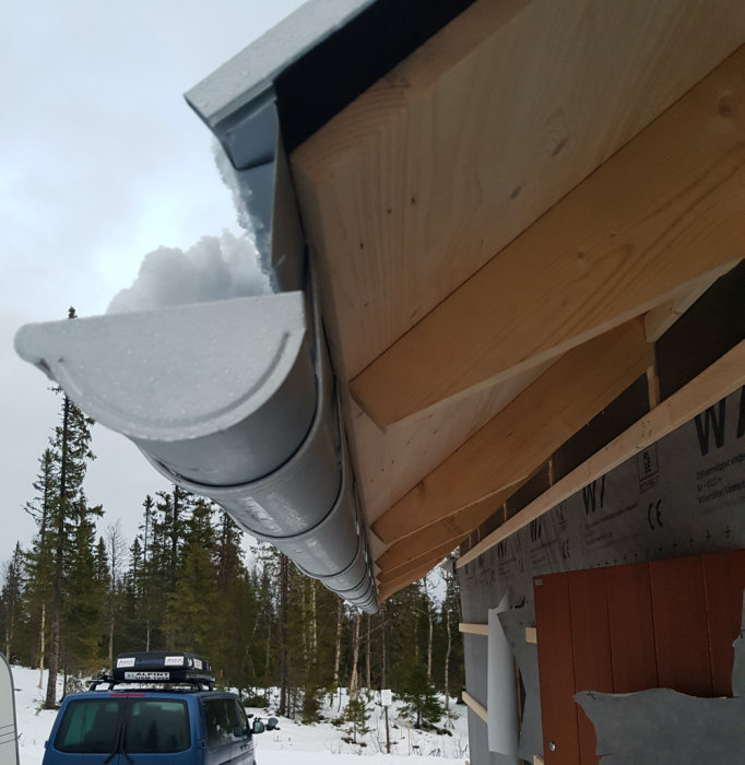 Hörnet av ett fritidshus med utstickande tak och hängränna som blockerar dörröppningen, omgiven av snöigt fjällandskap.