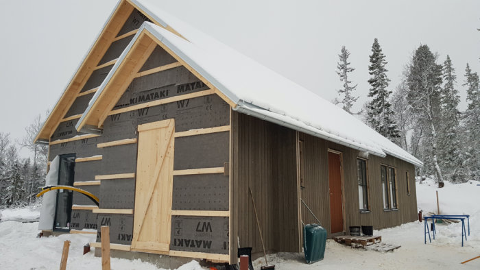 Fritidshus under konstruktion i snöigt landskap med utstickande tak och problematisk rännkrok vid entrédörren.