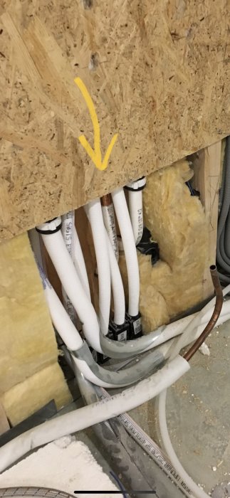 Vit VVS-rörinstallation med övergångar som passerar genom en isolerad vägg med en gul pil som indikerar riktning.