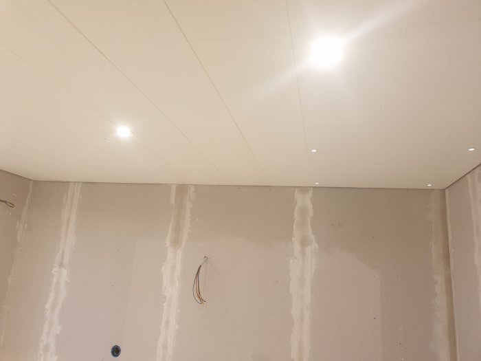 Nyinstallerade spotlights i ett vitmålat innertak med oslipade gipsskivor på väggen i ett badrum under renovering.