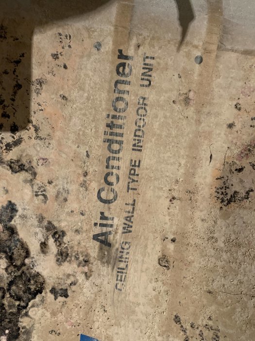 Delvis smutsigt pappfördral med texten 'Air Conditioner Ceiling/Wall Type Indoor Unit' som indikerar att det tillhör en AC-enhet.