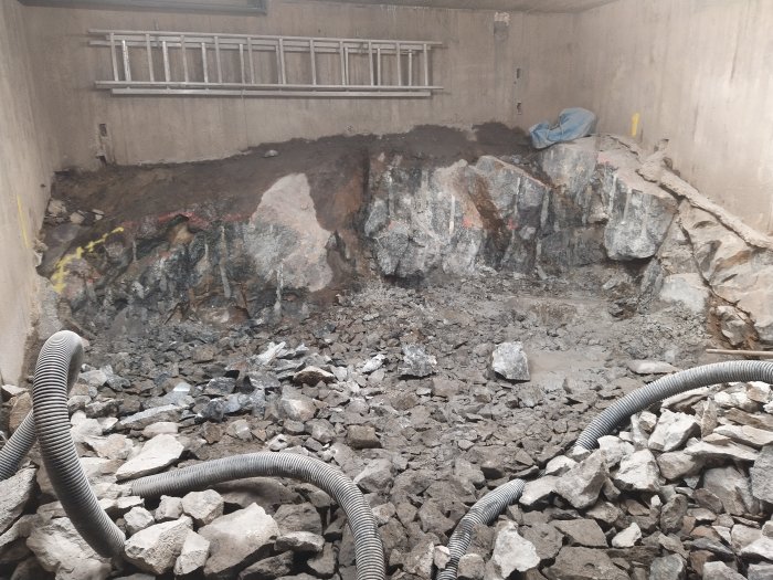 En grov uppbruten stenig mark med större stenblock och grus under konstruktion, omgiven av betongväggar.