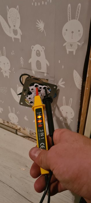 En hand håller en spänningsprovare mot en oskyddad elinstallation med tecknat djurmönster i bakgrunden.