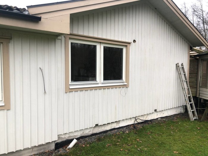 Dräneringsslang sticker upp ur marken vid husvägg med grus och gräsmatta, stege lutad mot vägg.