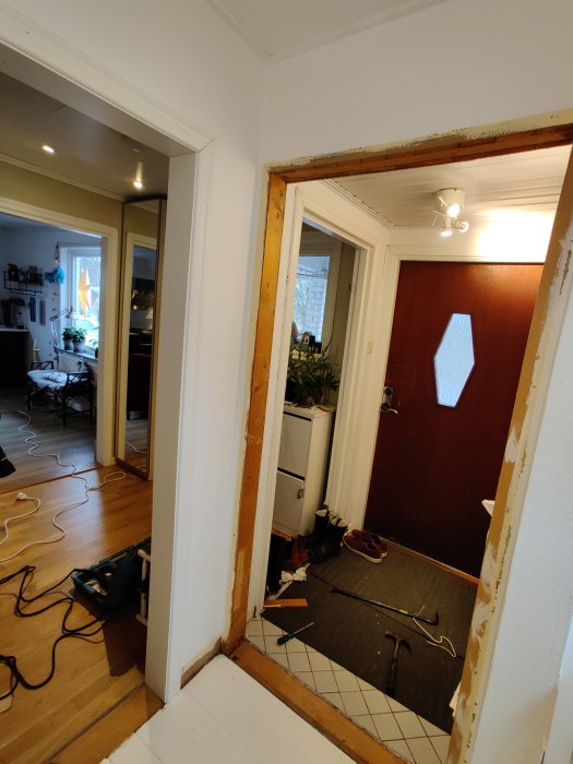 Åbning i væg, hvor en ramme er fjernet. Værktøj som hammer og skruetrækker er på gulvet. Dør til højre; stuevej med trægulv til venstre.