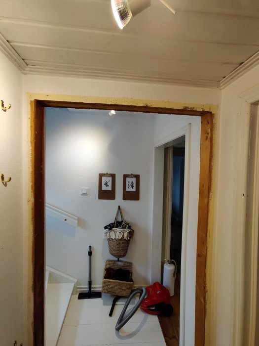 Åbning i væg, hvor dørkarmen er fjernet. Støvsuger og kurve på gulvet. To billeder hænger på den fjerneste væg. Lysekrone på loftet.