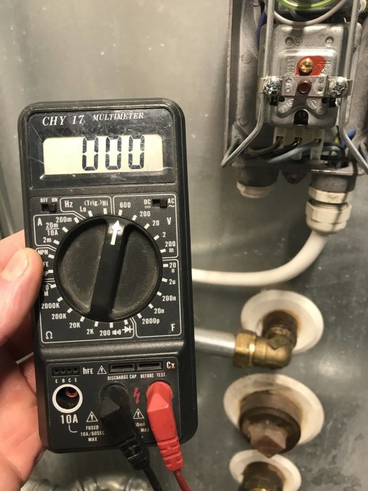 Multimeter visas med displayen 0.000 i framför en öppen elektrisk panel med kablar och anslutningar.