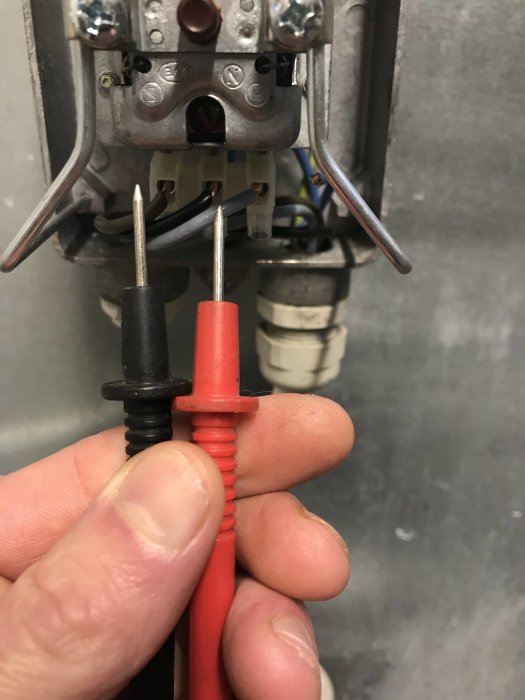 En person håller en svart och en röd multimeterprobe mot anslutningar märkta L1 och L2 i en elektrisk panel.