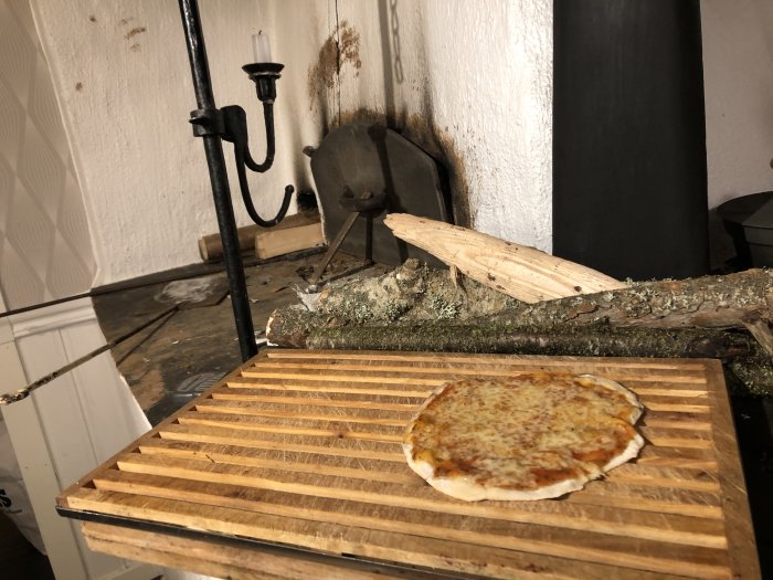 Nygräddad pizza på träspade framför en vedeldad stenugn med sotiga väggar och vedträn.