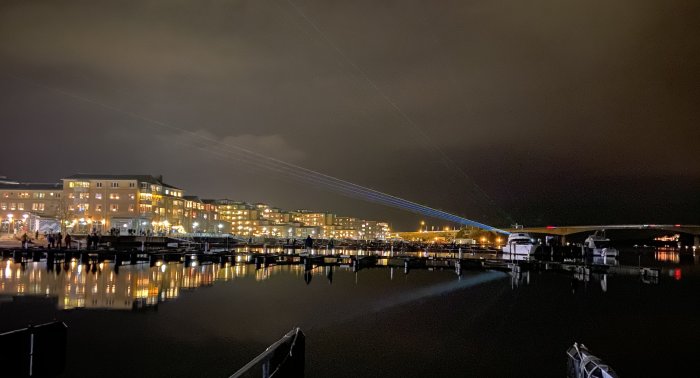 Kvällsbild över marina med belysta byggnader och båtar, speglade i vattnet, och strålkastarljus över himlen.