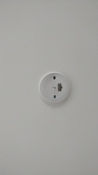 Plastlock med krok över eluttag på vit vägg, tänkt för upphängning av lampa.