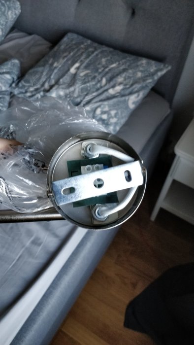 Hand som håller en lampa med metallbygel och kabel, avsedd för takmontering, framför en soffa.