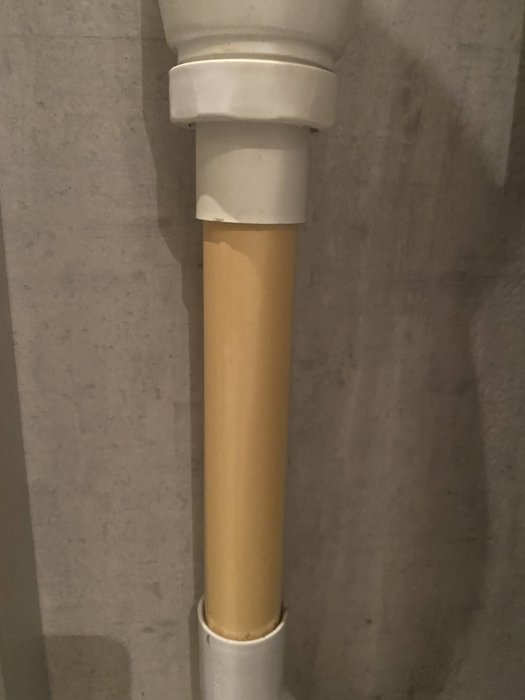 Ett avloppsrör i plast som är gulnat i mitten med vita kopplingar i båda ändarna mot en grå vägg.