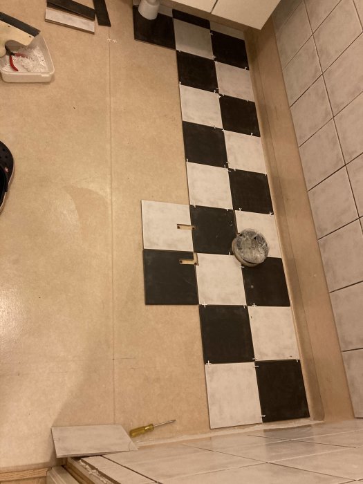Pågående kakelläggning med ett schackmönster på ett golv, med verktyg och lösa plattor runtomkring.