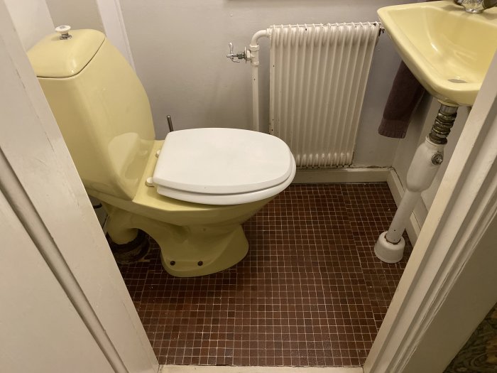 Liten toalett med gul WC och handfat samt ett vitt element på en brun mosaikgolv.