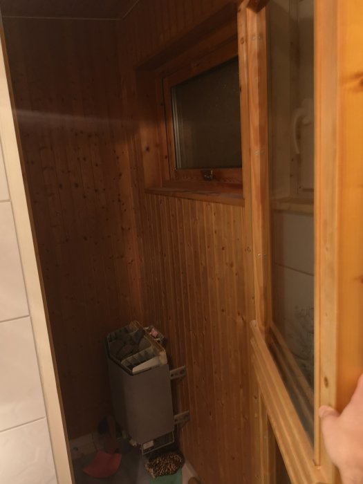 Trång bastu med träväggar och en liten fönster intill ett helkaklat badrum som ska renoveras.