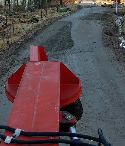 Röd schaktblad bakom en traktor på en ojämn grusväg som behöver underhåll.