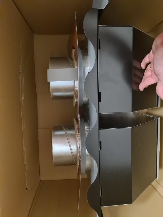 Ventilationsdelar och en hand som justerar en svart metallkanal i en kartong.