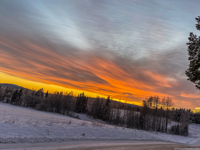 Vinterlandskap vid solnedgång med snötäckt mark och dramatiskt färgad himmel.