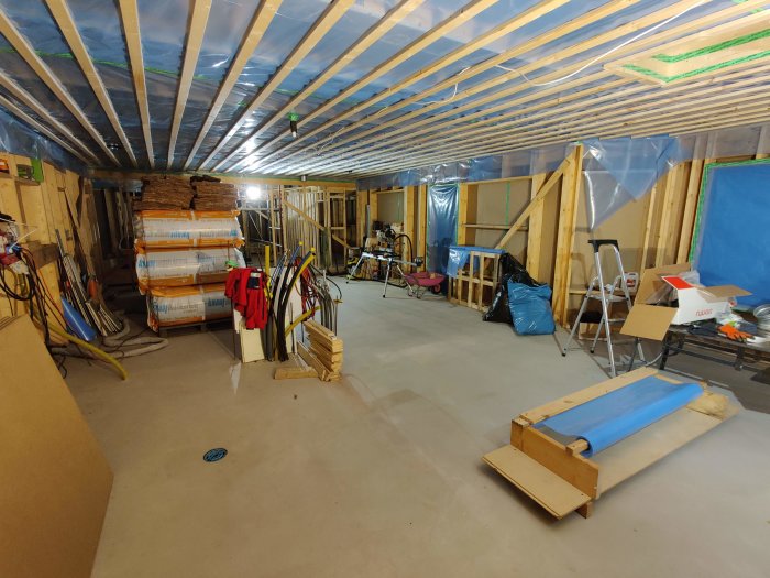 Renoveringsarbete i ett oisolerat rum med synliga takbjälkar, plast på väggarna och byggmaterial.