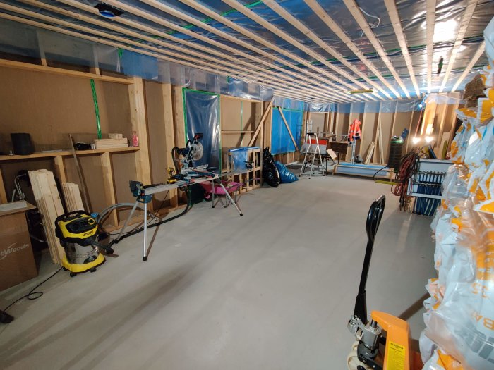 Oisolerat loft under renovering med synliga träreglar, plastklädd vägg, verktyg och avfuktare.