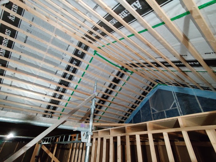 Innertak med synliga takstolar och isoleringsplast, samt ventilationsrör på en byggarbetsplats.