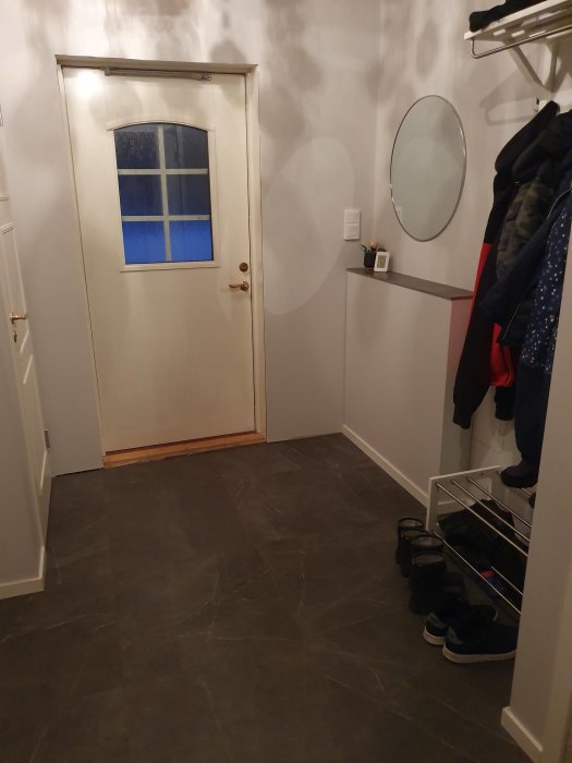 En hall med vit dörr, skoställ, spegel och klädhängare, grått golv och beige väggar.