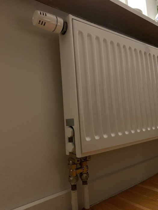 Nyinstallerad vit radiator med termostat och anslutningsrör på en vägg.