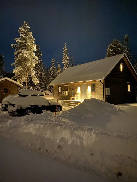 Belyst stuga på vintern med snötäckt landskap och parkerad bil.