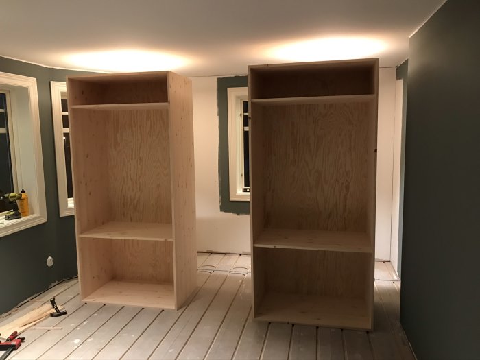 Två outbyggda garderobsstommar av furu i ett rum under renovering, verktyg syns på golvet.