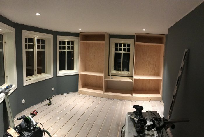 Nybyggda furugarderober med plywoodfyllning placerade mellan fönster i ett rum under renovering.