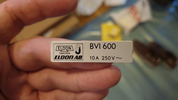 Etikett för ELDON AB BVI 600 strömbrytare, 10A 250V, hålls av en person, suddig bakgrund.